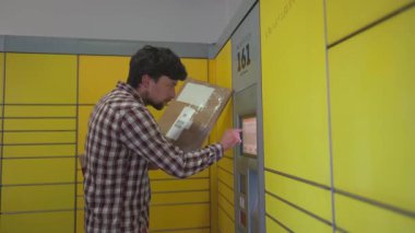 Sarı self servis posta terminali. Paket yollayan adam. Paket toplama noktası, bağlantısız self servis terminali, paket teslimatı. Kutuyu paket makinesine koyan adam. Paket depolama için depozito
