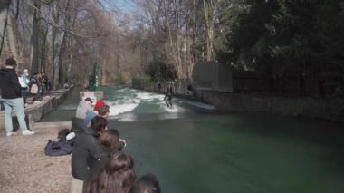 25 Mart 2022. Münih, Almanya. Sörfçüler bahar güneşli havalarda İngiliz Bahçelerinde bir Isar Nehri 'nde sörf yapıyor ve insanlar izliyor. Eisbach 'taki nehir sörfçüsü. Eisbachwelle am Englischen Bahçesi. Sörf Noktası