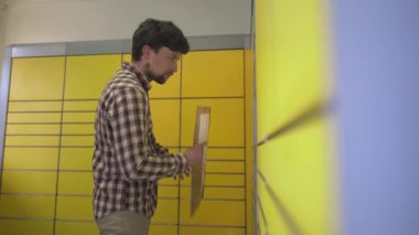 Sarı self servis posta terminali. Paket yollayan adam. Paket toplama noktası, bağlantısız self servis terminali, paket teslimatı. Kutuyu paket makinesine koyan adam. Paket depolama için depozito