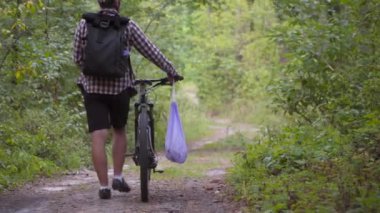 Bisiklet ve çöp torbasıyla ormanda yürüyen erkek bisikletçi spor yaparken çöp ve plastik topluyor. Bisikletçi çöp torbasında çöp topladı ve ormanda yürüdü. Bilinç. 