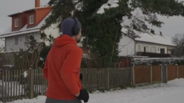 Erkek koşucu kışın Avrupa 'daki karlı bir parkta koşuyor ve büyük kulaklıklardan arka ve yan görüntülerden müzik dinliyor. Karlı parkta koşan bir adam ve büyük kulaklıklarla müzik dinliyor.. 
