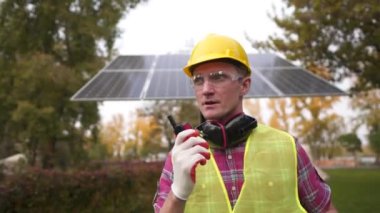 Erkek enerji sistemleri mühendisi kamerada yürüyor ve dışarıda güneş panelinin önünde telsizle konuşuyor. İşçi güneş enerjisi santralindeki koruyucu giysili meslektaşlarıyla iletişim kurmak için radyo kullanıyor.. 