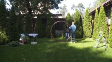 Erkek ve kadın, küçük işletme düğün dekoratörleri, açık hava bahçe töreni için ahşap bir çiçek kemeri kurun. Birlikte çalışıyorlar, güzel bir ortam yaratmak için detaylara çok dikkat ediyorlar.. 