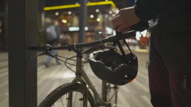 Bisikletli erkek bisikletçi bisikletini direğe park etti ve şehirde akşam üstü bisikletinin gidonuna kırmızı ışıkla kaskını astı. Bisikletçiler için yol güvenliği teması. Bisiklet için kırmızı ışıklı kask. 