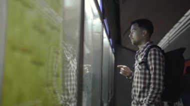 Adam Almanya 'daki toplu taşıma istasyonunda arka plandaki bilgilere bakıyor, akıllı telefonundan detayları kontrol ediyor. Yolcu bilgi panosundaki metro tren tarifesine bakıyor.. 