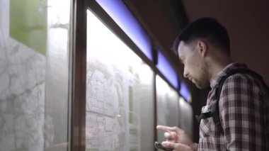 Adam Almanya 'daki toplu taşıma istasyonunda arka plandaki bilgilere bakıyor, akıllı telefonundan detayları kontrol ediyor. Yolcu bilgi panosundaki metro tren tarifesine bakıyor.. 