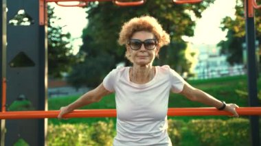 Olgun Avrupalı kadın yazın dışarıda egzersiz yapmaktan hoşlanıyor. Açık hava spor salonundaki barfiks çubuklarını kullanarak sırtını esnetmeye ve ısıtmaya odaklanıyor. Sağlıklı yaşam tarzı, zindelik ve yaşlılık. 