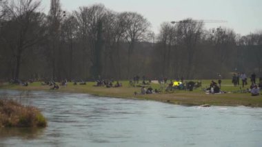 22 Mart 2022. Münih, Almanya. İnsanlar baharda güneşli ve sıcak havalarda Englischer Bahçesi 'ndeki Isar Nehri yakınlarındaki yeşil çimlerde dinleniyorlar. İnsanlar sıcak günün tadını çıkarıyorlar, Münih 'teki Isar Nehri yakınlarında güneşleniyorlar.. 