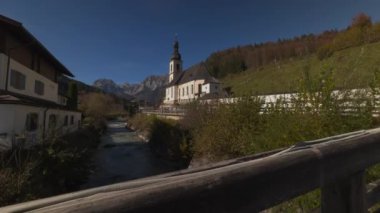 Ramsau 'daki St. Sebastian Kilisesi, Bavyera Almanya, sonbahar dağlarına karşı kurulmuş, güneşli gökyüzünün altındaki nehirler. Bu ikonik Alman simgesi, gezginleri ve doğa aşıklarını cezbeden, pitoresk bir manzara sunuyor.. 