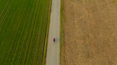 Asfalt yolda bisiklet süren kadın iki yeşil ve sarı hava manzaralı alanı ayırıyor. İlkbahar insansız hava aracı manzaralı tarlalarda bisiklet sürmek. İki renkli tarla yolu ile bölünmüş.. 
