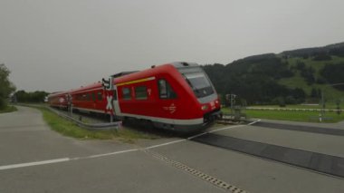 Almanya, Bavyera 'da demiryolu geçidi, yanıp sönen ışıklar ve kırmızı bir bölgesel tren hızıyla kapanan bir kapıyla. Bisikletçiler, dikkat ve güvenliğin önemini vurgulayarak bir bisiklet yolunun yakınından geçerler.. 