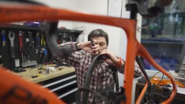 Erkek bisiklet tamircisi, profesyonel tamir atölyesinde yüksek performanslı otoyol bisikletinin lastiğini dikkatlice değiştiriyor. Etrafı aletlerle, yedek parçalarla ve duvarlarda düzenli ekipmanlarla çevrili.. 