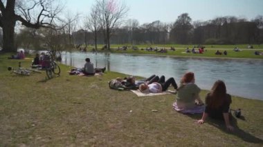 22 Mart 2022. Münih, Almanya. İnsanlar baharda güneşli ve sıcak havalarda Englischer Bahçesi 'ndeki Isar Nehri yakınlarındaki yeşil çimlerde dinleniyorlar. İnsanlar sıcak günün tadını çıkarıyorlar, Münih 'teki Isar Nehri yakınlarında güneşleniyorlar.. 