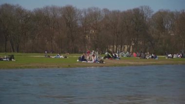 22 Mart 2022. Münih, Almanya. İnsanlar baharda güneşli ve sıcak havalarda Englischer Bahçesi 'ndeki Isar Nehri yakınlarındaki yeşil çimlerde dinleniyorlar. İnsanlar sıcak günün tadını çıkarıyorlar, Münih 'teki Isar Nehri yakınlarında güneşleniyorlar.. 