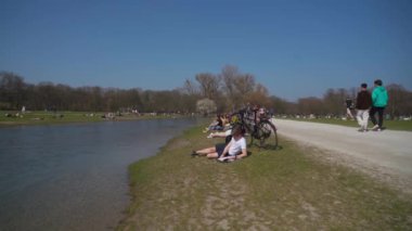 22 Mart 2022. Münih, Almanya. İnsanlar baharda güneşli ve sıcak havalarda Englischer Bahçesi 'ndeki Isar Nehri yakınlarındaki yeşil çimlerde dinleniyorlar. İnsanlar sıcak günün tadını çıkarıyorlar, Münih 'teki Isar Nehri yakınlarında güneşleniyorlar.. 