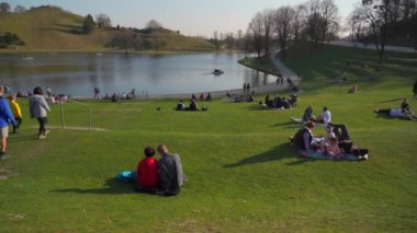 25 Mart 2022. Münih, Almanya. İnsanlar sıcak ve güneşli bahar havasında Münih 'teki Olimpik Park' ın yanındaki yeşil çimlerde dinleniyorlar. Olympiapark Münih. Olympiaberg ve Olympiasee in Olympiapark. 
