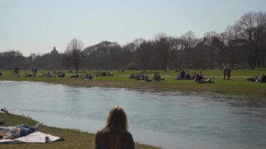 22 Mart 2022. Münih, Almanya. İnsanlar baharda güneşli ve sıcak havalarda Englischer Bahçesi 'ndeki Isar Nehri yakınlarındaki yeşil çimlerde dinleniyorlar. İnsanlar sıcak günün tadını çıkarıyorlar, Münih 'teki Isar Nehri yakınlarında güneşleniyorlar.. 