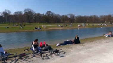 22 Mart 2022. Münih, Almanya. İnsanlar baharda güneşli ve sıcak havalarda Englischer Bahçesi 'ndeki Isar Nehri yakınlarındaki yeşil çimlerde dinleniyorlar. İnsanlar sıcak günün tadını çıkarıyorlar, Münih 'teki Isar Nehri yakınlarında güneşleniyorlar.. 