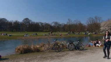 22 Mart 2022. Münih, Almanya. İnsanlar baharda güneşli ve sıcak havalarda Englischer Bahçesi 'ndeki Isar Nehri yakınlarındaki yeşil çimlerde dinleniyorlar. İnsanlar sıcak günün tadını çıkarıyorlar, Münih 'teki Isar Nehri yakınlarında güneşleniyorlar.. 