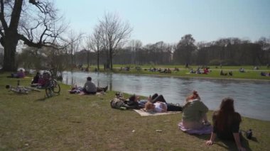 22 Mart 2022. Münih, Almanya. İnsanlar baharda güneşli ve sıcak havalarda Englischer Bahçesi 'ndeki Isar Nehri yakınlarındaki yeşil çimlerde dinleniyorlar. İnsanlar sıcak günün tadını çıkarıyorlar, Münih 'teki Isar Nehri yakınlarında güneşleniyorlar.. 