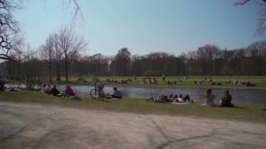 22 Mart 2022. Münih, Almanya. İnsanlar baharda güneşli ve sıcak havalarda Englischer Bahçesi 'ndeki Isar Nehri yakınlarındaki yeşil çimlerde dinleniyorlar. İnsanlar sıcak günün tadını çıkarıyorlar, Münih 'teki Isar Nehri yakınlarında güneşleniyorlar.. 