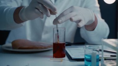 Beyaz önlüklü bir bilim adamı laboratuarda şırınga kullanarak bir parça tavuk etine kırmızı sıvı enjekte ediyor. Laboratuvar ekipmanlarıyla çevrili deney, GDO araştırmalarına odaklanıyor..