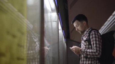 Adam Almanya 'daki toplu taşıma istasyonunda arka plandaki bilgilere bakıyor, akıllı telefonundan detayları kontrol ediyor. Yolcu bilgi panosundaki metro tren tarifesine bakıyor.. 
