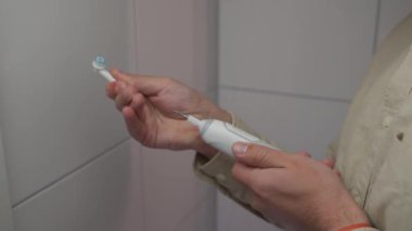 Banyoda beyaz elektrikli diş fırçası tutan, mod değiştiren ve fırça başlığını değiştiren tişörtlü adamın yakın çekimi. Erkeklerin elleri ultrasonik diş fırçası kullanıyor, başlarını değiştiriyor. Diş hijyeni.. 