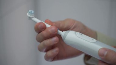Banyoda beyaz elektrikli diş fırçası tutan, mod değiştiren ve fırça başlığını değiştiren tişörtlü adamın yakın çekimi. Erkeklerin elleri ultrasonik diş fırçası kullanıyor, başlarını değiştiriyor. Diş hijyeni.. 