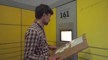 Sarı self servis posta terminali. Paket yollayan adam. Paket toplama noktası, bağlantısız self servis terminali, paket teslimatı. Kutuyu paket makinesine koyan adam. Paket depolama için depozito
