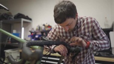 Bisiklet tamircisi tamircisinde çalışıyor, çakıllı bir bisiklete gidon bandı takıyor. Teknisyen bandı ustalıkla gidonun etrafına sarar, atölyede hassasiyet ve kaliteye odaklanır.. 