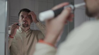Aynadaki yansımalar, işten önce gömlekli bir adam dişlerini siyah ultrasonik elektrikli diş fırçasıyla fırçaladı, kendini beğendi, saçını beğendi ve aynada kendine bakarken onu düzeltti.. 
