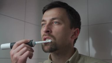 Bir adam işe hazırlanırken banyoda elektrikli diş fırçasıyla dişlerini fırçalıyor. Sabah rutinini takip ediyor, kişisel hijyen, tımar ve oral sağlığa odaklanıyor.