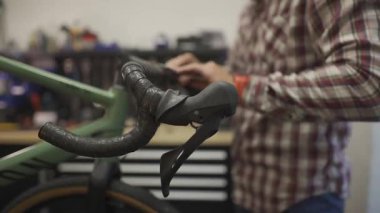 Bisiklet tamircisi tamircisinde çalışıyor, çakıllı bir bisiklete gidon bandı takıyor. Teknisyen bandı ustalıkla gidonun etrafına sarar, atölyede hassasiyet ve kaliteye odaklanır.. 