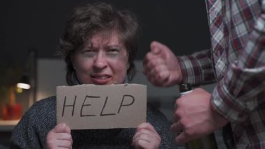 Şiddet korkusu olan yaralı bir kadın yardım için yalvaran yardım işaretini tutuyor. Yüzünde çürükler ve kırık diş de dahil olmak üzere yaralar var. Erkek yumruklarını sıkması, aile içi şiddet ve korkuyu yansıtıyor.