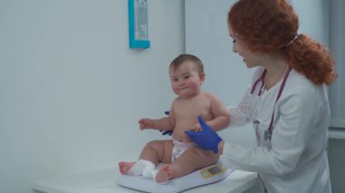 Çocuk doktoru rutin kontroller sırasında bebeği tartar. Bebek tıbbi ölçekte otururken doktor eldiven ve steteskop takıyor, çocukların sağlığını izliyor. Klinikte standart bir pediatrik muayene.. 