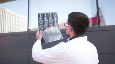 Beyaz önlüklü ve gözlüklü genç bir doktor hastanenin önünde duruyor. Tomografiyi inceliyor. Kısa bir sigara molası vererek, sınırlı zamanına rağmen tıbbi görüntüleri analiz ederek işine odaklanıyor.