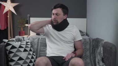 Evdeki kanepede oturan bir adam, acı içinde boynunu tutuyor. Boyun desteği için siyah bir yaka takıyor, boyun yaralanması ya da gerilme rahatsızlığı gösteriyor, rahatlama ve iyileşme arıyor.. 