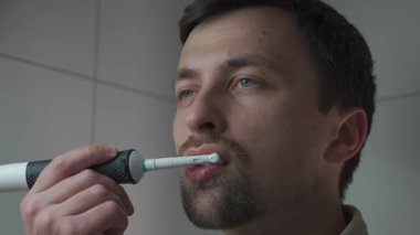 Bir adam sabah rutini sırasında banyoda elektrikli diş fırçasıyla dişlerini fırçalıyor. İşe hazırlanıyor, kişisel hijyenine, bakımına ve ağız sağlığına özen gösteriyor.. 