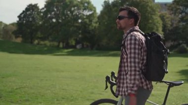Ekose gömlekli ve siyah sırt çantalı genç bir bisikletçi çakıllı bisikletini Almanya 'daki güneşli bir parkta yürüyor. Anın tadını çıkarıyor, etrafa bakıyor, rahatlıyor ve yaz manzarasını seyrediyor.