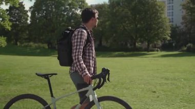Ekose gömlekli ve siyah sırt çantalı genç bir adam Almanya 'da güneşli bir parkta çakıllı bisikletinin yanında yürüyor, anın tadını çıkarıyor, etrafına bakıyor ve huzurlu yaz atmosferinde ıslanıyor.