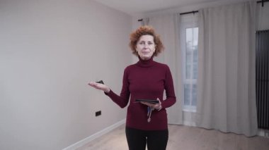 Olgun bir emlakçı kadın müşterilere boş bir daire sunar. Anahtarları elinde tutuyor, modern, aydınlık bir yerde duruyor. Profesyonel ve kendine güveni tam, mülkü satılık veya kiralık olarak gösteriyor