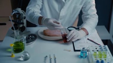 Laboratuvardaki bilim adamı çiğ tavuk eti üzerinde bir deney yapıyor. Eldiven ve laboratuvar önlüğü giyip şırıngadan filetoya sıvı enjekte etmiş. Laboratuvar ekipmanları, test tüpleri ve mikroskop etrafını sarsın.