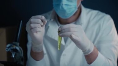 Koruyucu ekipmanlı erkek laboratuvar teknisyeni bilimsel bir deney için şırınga kullanarak test tüpünden yeşil sıvı çıkarıyor. Laboratuvar ortamında bir mikroskop bulunmakta, araştırma ve analizlerin vurgulanması. 