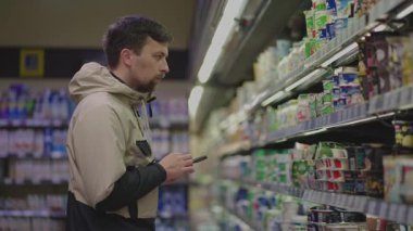 Süpermarkette alışveriş yapan bir adam, ürünlerini seçerken market listesini akıllı telefondan kontrol eder. Gıda etiketlerini okuyor, beslenme gerçeklerini inceliyor ve sağlıklı bir seçim yapmak için gerekli malzemeleri karşılaştırıyor.