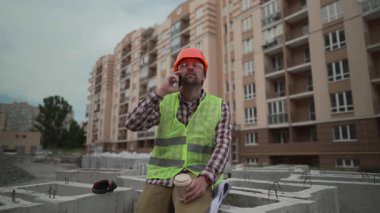 İnşaat mühendisi, inşaat alanındaki beton yapıların üzerinde duruyor. Kasketli ve yansıtıcı yelek giyiyor. Kahve içiyor, telefonda konuşuyor ve site incelenirken planlar, belgeler tutuyor.. 