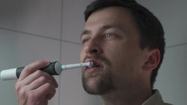 Bir adam sabah rutininin bir parçası olarak banyoda elektrikli diş fırçası kullanıyor. İşe hazırlanıyor, kişisel hijyene odaklanıyor, tımar ediyor ve ağız sağlığını koruyor.