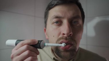 Genç adam sabah işe gitmeden önce elektrikli diş fırçasıyla dişlerini fırçalar ve kameraya geniş açıdan bakar. Diş fırçalama seansı. Sabah hijyeni. Günlük diş fırçalama rutini. Ağız temizliği.. 
