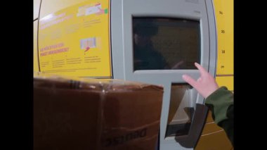 Kadının Almanya 'daki DHL otomatik servis dolap istasyonu üzerinden paket gönderirken ki görüntüsü. Dokunmatik ekranı kullanan ve paketi güneşli bir günde sarı teslimat kutusuna koyan eller.. 