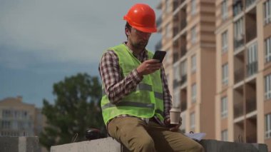 Sert şapkalı ve yansıtıcı yelekli erkek inşaat mühendisi kahve içerek gökdelen şantiyesinde güçlendirilmiş beton yapıda akıllı telefon kullanıyor. Güneşli hava, şehir silüeti